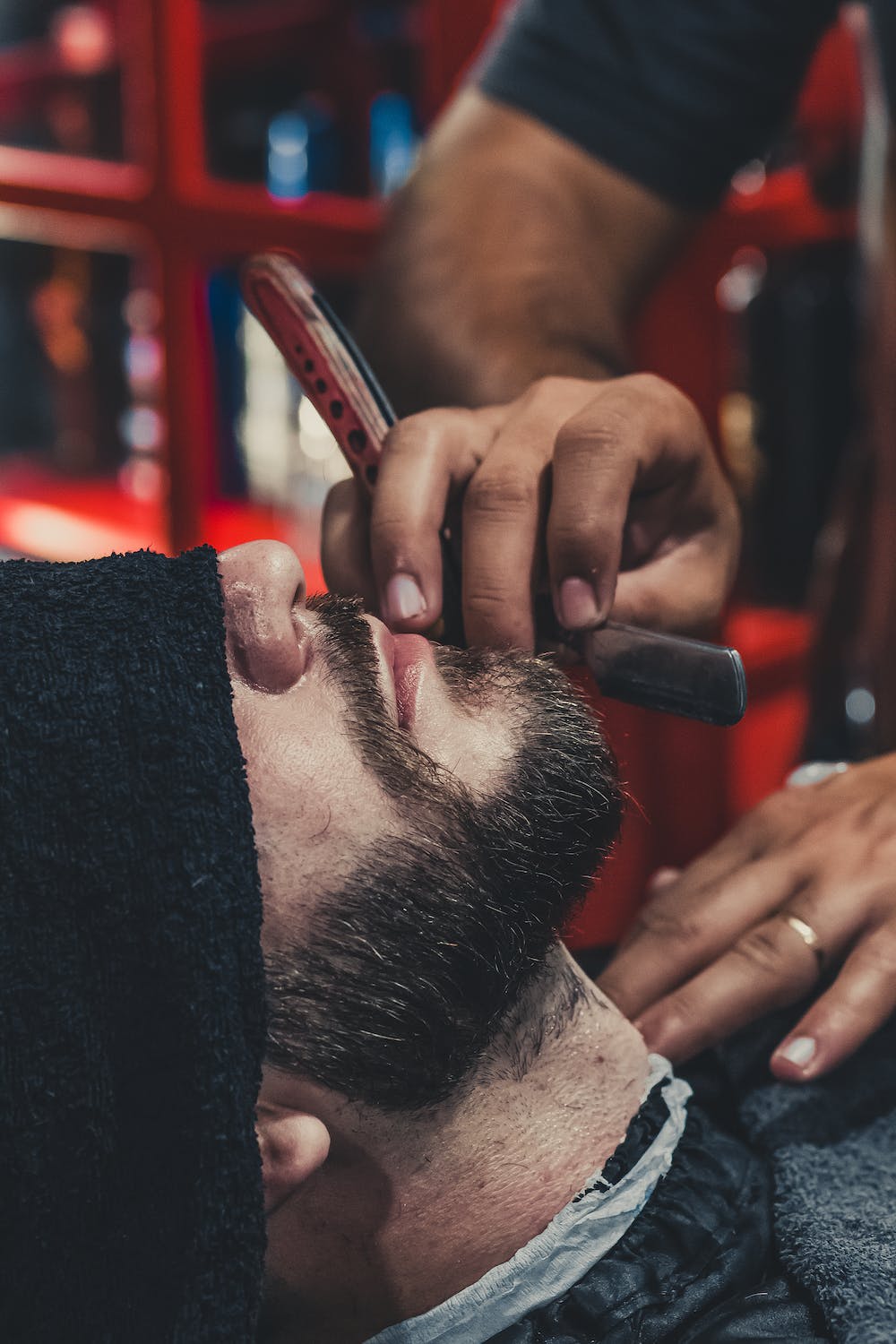 Barber straight razor shaving customers neck area while customer relaxes with a warm towel over his eyes